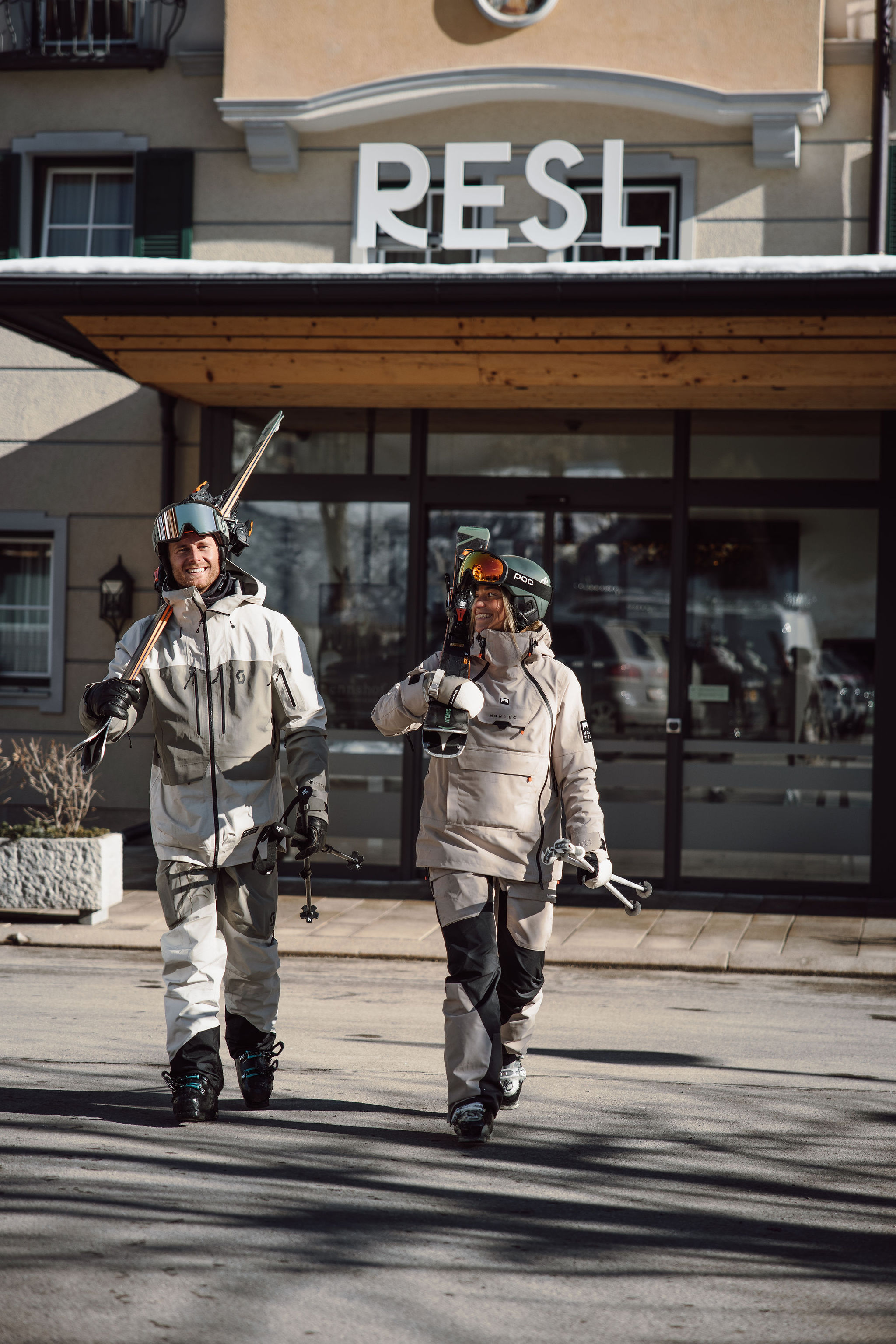 Zwei Personen in Skiausrüstung gehen lachend mit Skiern vor einem Gebäude
